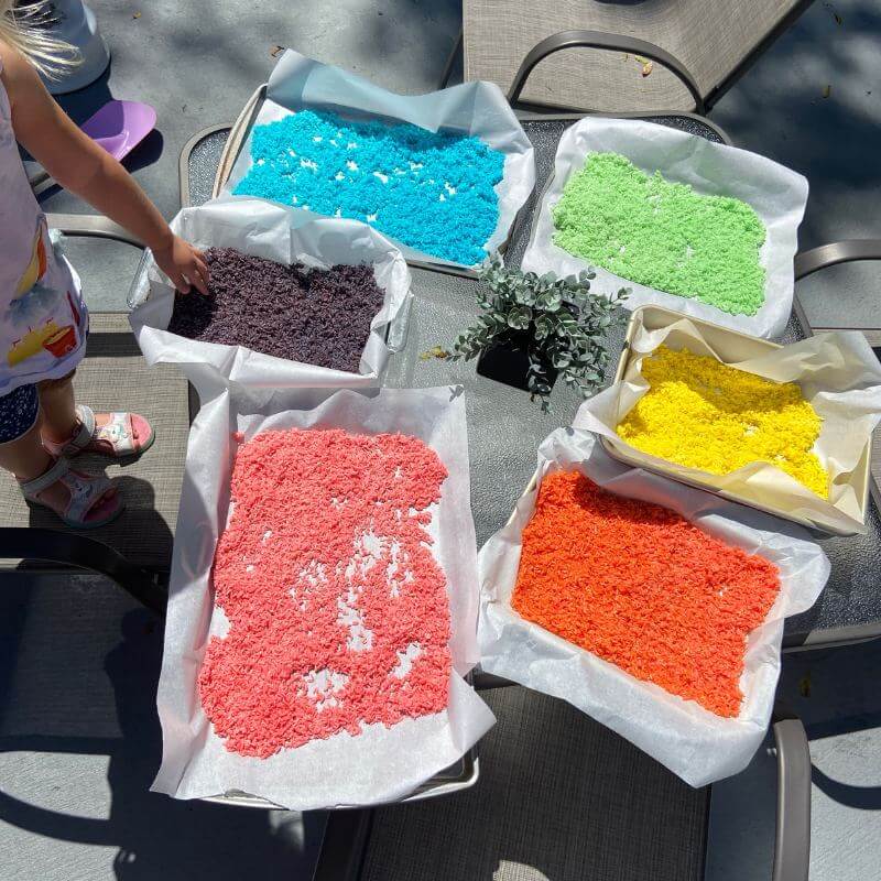 six different colors of dyed rice spread out on baking sheets to dry